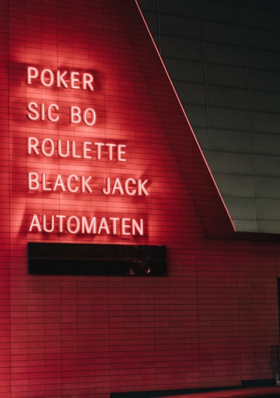 Red neon letters are mounted on a dark tiled wall, displaying the words Poker, Sic Bo, Roulette, Black Jack, and Automaten.