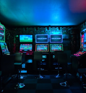 Dimly lit gaming arcade featuring a variety of brightly illuminated slot machines and video gaming screens. Neon signs and colorful displays create a vibrant atmosphere. The arrangement of gaming machines suggests an inviting space for entertainment, with chairs positioned for players.