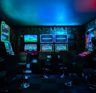 Dimly lit gaming arcade featuring a variety of brightly illuminated slot machines and video gaming screens. Neon signs and colorful displays create a vibrant atmosphere. The arrangement of gaming machines suggests an inviting space for entertainment, with chairs positioned for players.