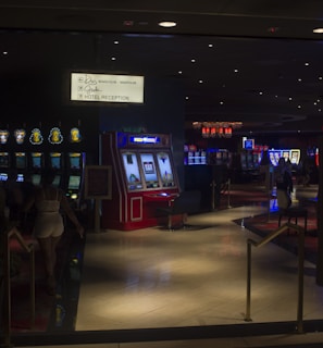 A dimly lit casino with several rows of slot machines, some displaying bright graphics and lights. There are a few people scattered throughout the area, engaging with the machines. The floor is reflective and polished, adding to the ambiance, while the ceiling is dotted with numerous small lights.