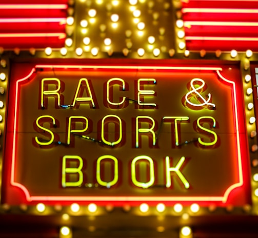 Bright neon lights on a marquee sign display the words 'RACE & SPORTS BOOK'. The sign is surrounded by rows of yellow and white lights against a red background, evoking a vibrant, energetic atmosphere typically associated with casinos or entertainment venues.