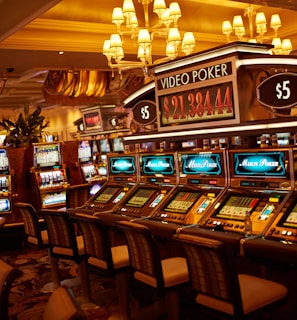 A casino floor featuring rows of brightly lit video poker machines. The area is illuminated by elegant chandeliers, with a display board showing a large sum of money, suggesting a possible jackpot. The rich decor includes luxurious carpets and comfortable seating, adding to the opulent atmosphere.