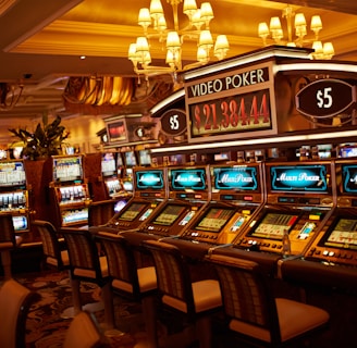 A casino floor featuring rows of brightly lit video poker machines. The area is illuminated by elegant chandeliers, with a display board showing a large sum of money, suggesting a possible jackpot. The rich decor includes luxurious carpets and comfortable seating, adding to the opulent atmosphere.