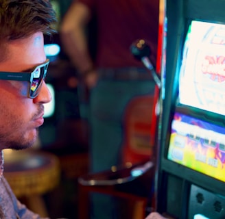 A man wearing sunglasses is intently focused on a slot machine, with colorful lights and graphics illuminating the screen. The scene is set indoors, likely in a casino or gaming area. His attire includes a patterned shirt and a blazer.