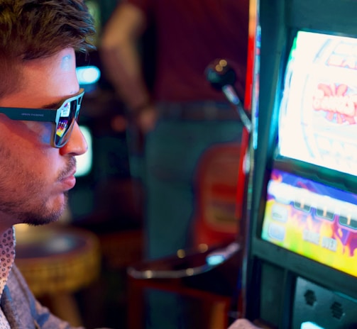 A man wearing sunglasses is intently focused on a slot machine, with colorful lights and graphics illuminating the screen. The scene is set indoors, likely in a casino or gaming area. His attire includes a patterned shirt and a blazer.
