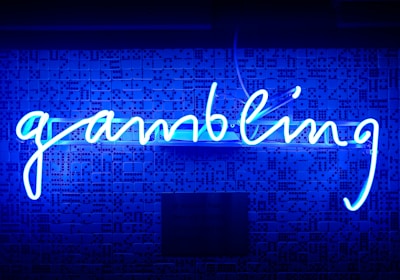 A bright blue neon sign spells out the word 'gambling' in cursive script. The background features a textured wall covered in domino tiles, also illuminated by the blue light, creating a vivid and visually striking scene.