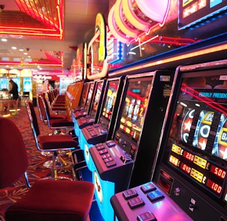 A brightly lit casino interior featuring a row of colorful slot machines. The neon lights create a vibrant atmosphere, with patterned carpeting adding to the decor. Stools are aligned neatly in front of each machine.