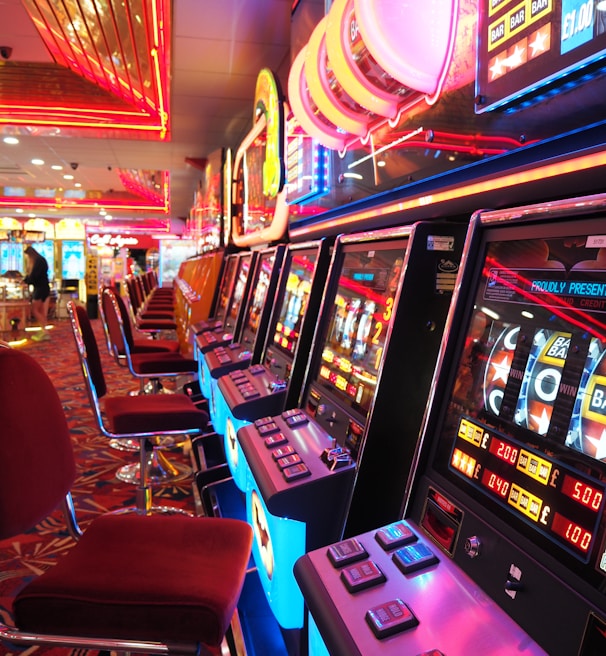 A brightly lit casino interior featuring a row of colorful slot machines. The neon lights create a vibrant atmosphere, with patterned carpeting adding to the decor. Stools are aligned neatly in front of each machine.