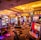 A vibrant casino interior filled with rows of colorful slot machines, each illuminated with bright lights and displays. The ceiling is adorned with elegant chandeliers and golden drapes, creating a luxurious atmosphere. The patterned carpet adds to the elaborate design of the space.