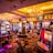 A vibrant casino interior filled with rows of colorful slot machines, each illuminated with bright lights and displays. The ceiling is adorned with elegant chandeliers and golden drapes, creating a luxurious atmosphere. The patterned carpet adds to the elaborate design of the space.