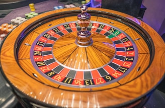 A roulette wheel with alternating red and black numbered pockets on a wooden platform. The wheel contains a small white ball resting in one of the pockets, indicating gameplay. Surrounding the wheel are stacks of colorful poker chips, suggesting a casino environment.