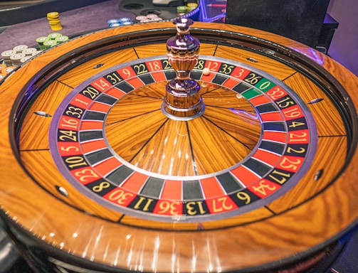 A roulette wheel with alternating red and black numbered pockets on a wooden platform. The wheel contains a small white ball resting in one of the pockets, indicating gameplay. Surrounding the wheel are stacks of colorful poker chips, suggesting a casino environment.