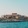 A seaside view of a large building labeled as a casino, situated on a rocky shore. The building is surrounded by palm trees and other smaller structures. The calm sea water reflects the sky, and there are several closed umbrellas lined along the waterfront.