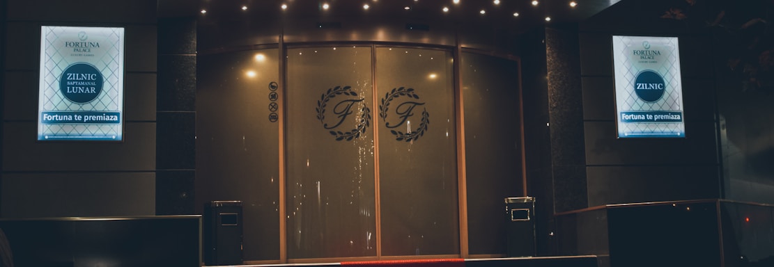 A neon-lit casino entrance with a red carpet leading up to large double doors. The sign reads 'Fortuna Palace Luxury Games' in bright red and white neon lights. The dimly lit atmosphere suggests a night-time setting. There are two digital displays on either side of the entrance showing promotional content.