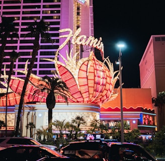 A brightly lit casino with neon lights in vibrant red, pink, and blue, featuring a large flamingo signage. Palm trees are visible in the foreground with vehicles passing by on the street at night.
