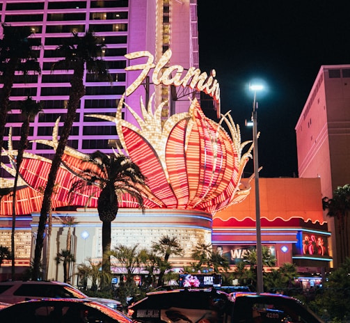 A brightly lit casino with neon lights in vibrant red, pink, and blue, featuring a large flamingo signage. Palm trees are visible in the foreground with vehicles passing by on the street at night.